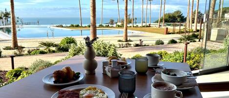 A breakfast table with plates of food, cups, and cutlery, overlooking a pool and palm trees.