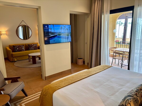Quarto de hotel com cama, sofá, televisão e varanda com mesa e cadeiras.