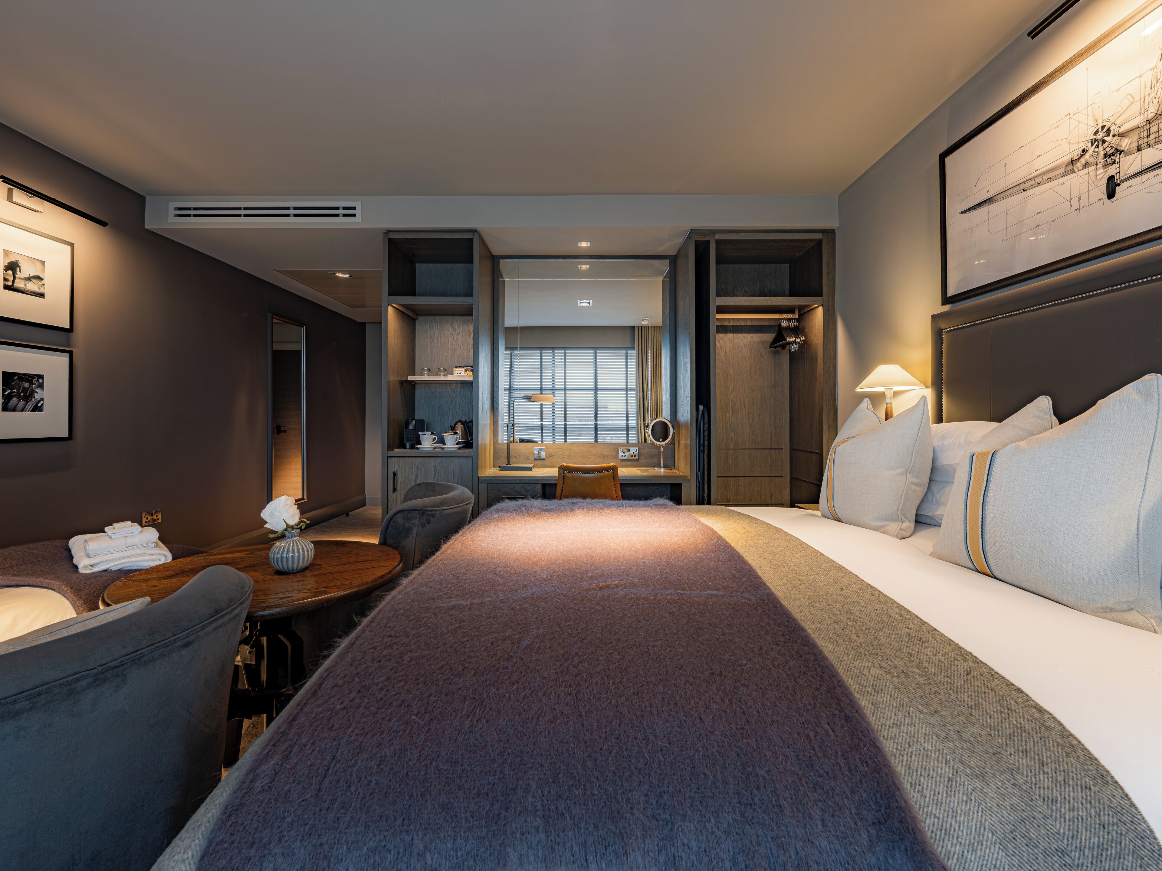 Una habitación de hotel con cama, escritorio, silla, una mesita con toallas, espejo y ventana con persianas.