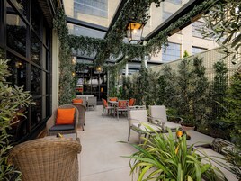 Un patio interior con mobiliario de mimbre, plantas en macetas y vista a la ciudad a través de amplias ventanas.