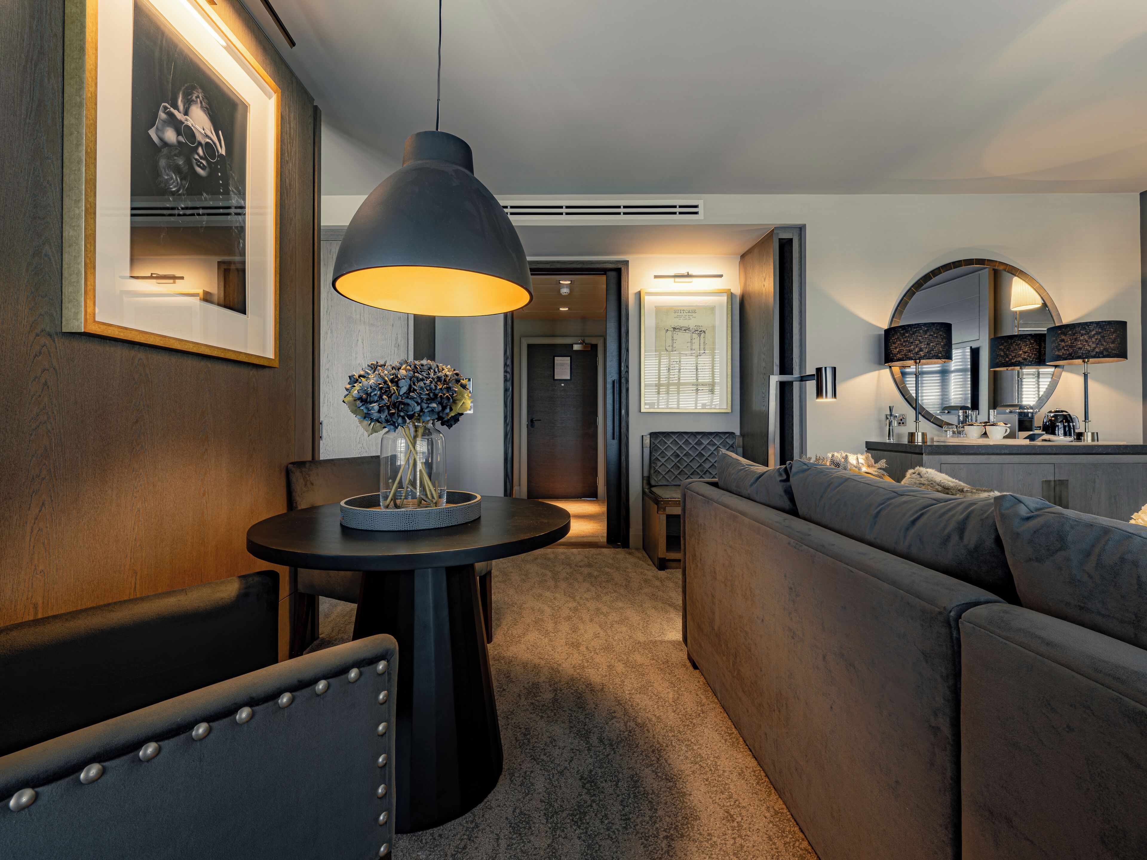 A modern hotel room with a round dining table, a sofa, a large pendant lamp, and a mirror above the sink.