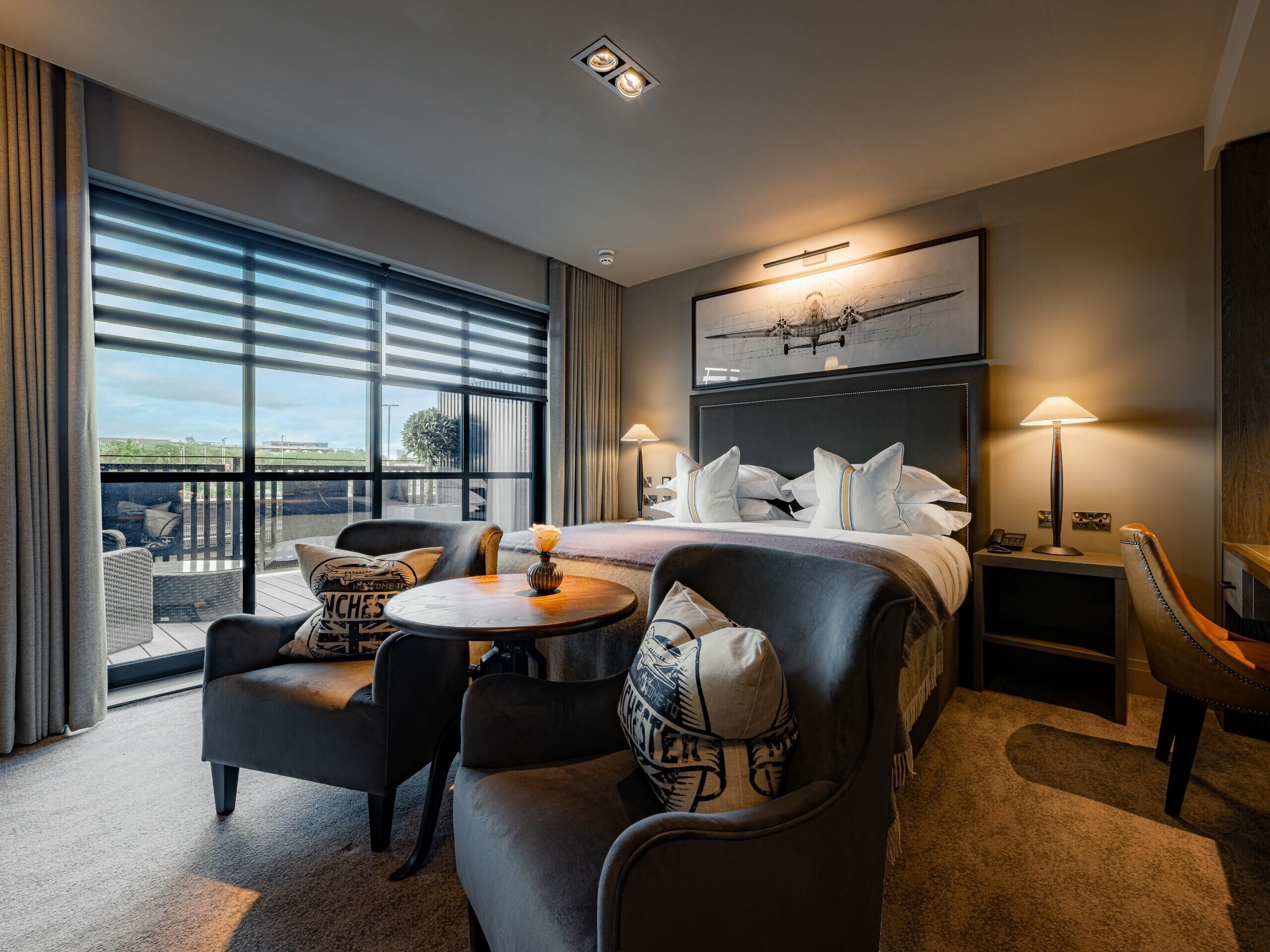 A hotel room with a large bed, two bedside tables, a round coffee table, a sofa, and a view of the outdoors through large windows.
