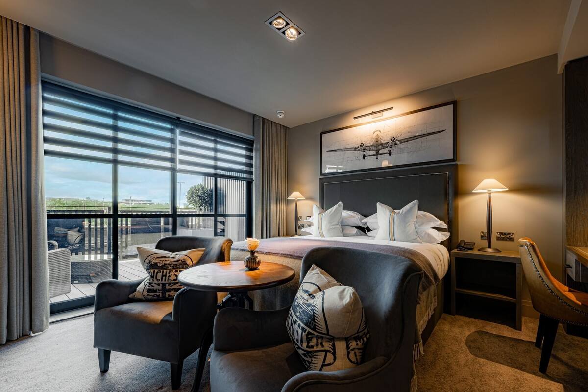 Habitación de hotel con una cama grande, dos mesitas de noche, una mesa de centro redonda, un sofá y vistas al exterior a través de amplias ventanas.