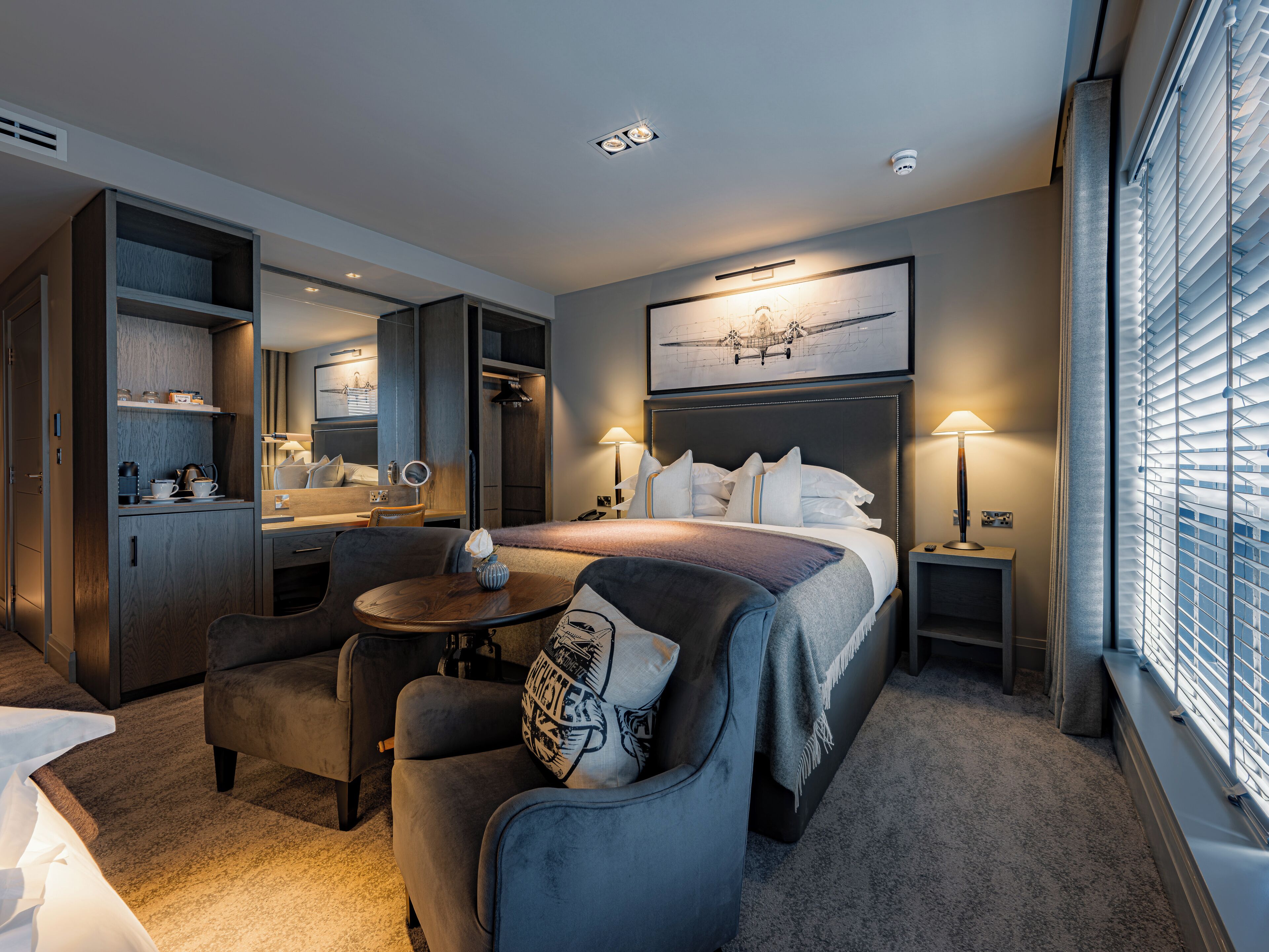 Ein Hotelzimmer mit Bett, Schreibtisch, Stuhl, einem kleinen Tisch, einem Spiegel und einem Fenster mit Jalousien.