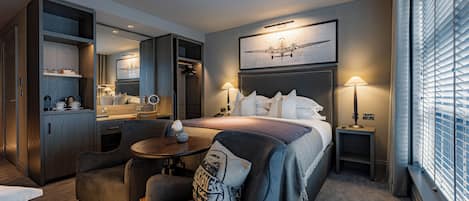 A hotel room with a bed, a desk, a chair, a small table, a mirror, and a window with blinds.
