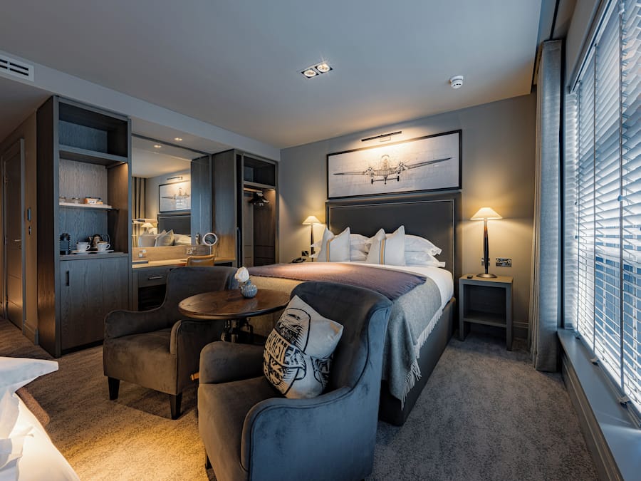 Habitación de hotel con cama, escritorio, silla, mesita, espejo y ventana con persianas.