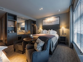 A hotel room with a bed, a desk, a chair, a small table, a mirror, and a window with blinds.