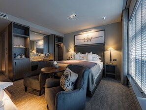 Habitación de hotel con cama, escritorio, silla, mesita, espejo y ventana con persianas.