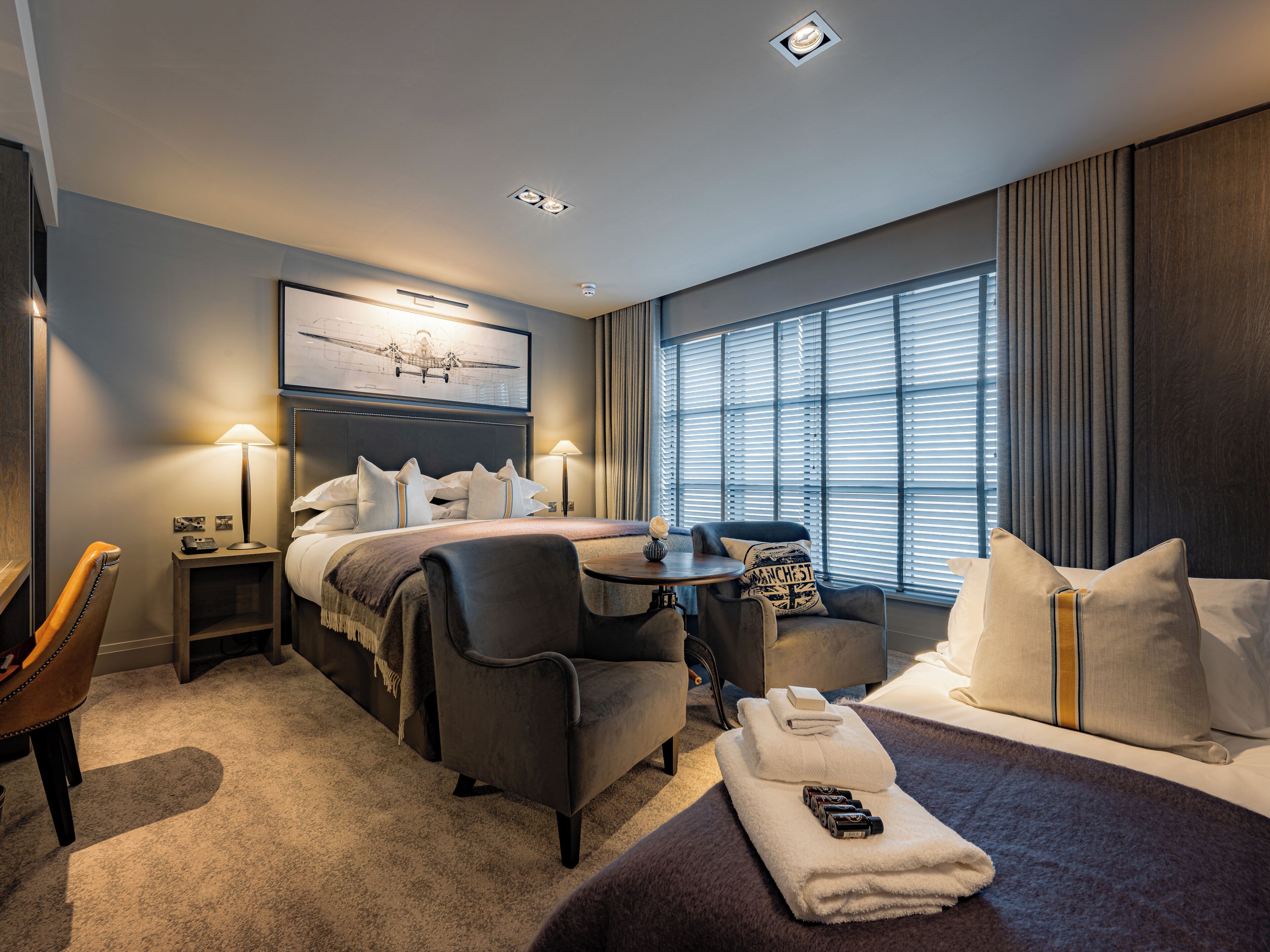 A hotel room with a bed, two armchairs, a small table, a desk, and a window with blinds.