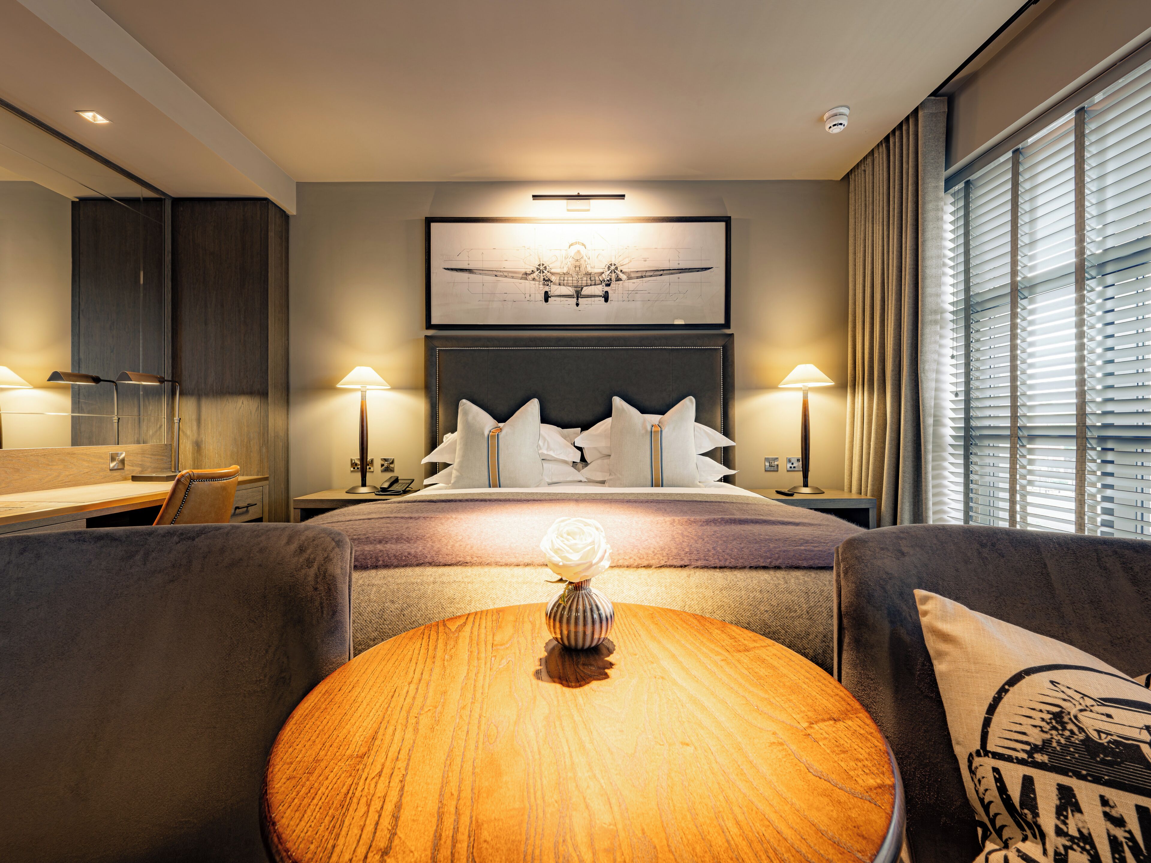 Une chambre d’hôtel avec un lit, des lampes de chevet, une table ronde en bois et une grande fenêtre avec des stores.