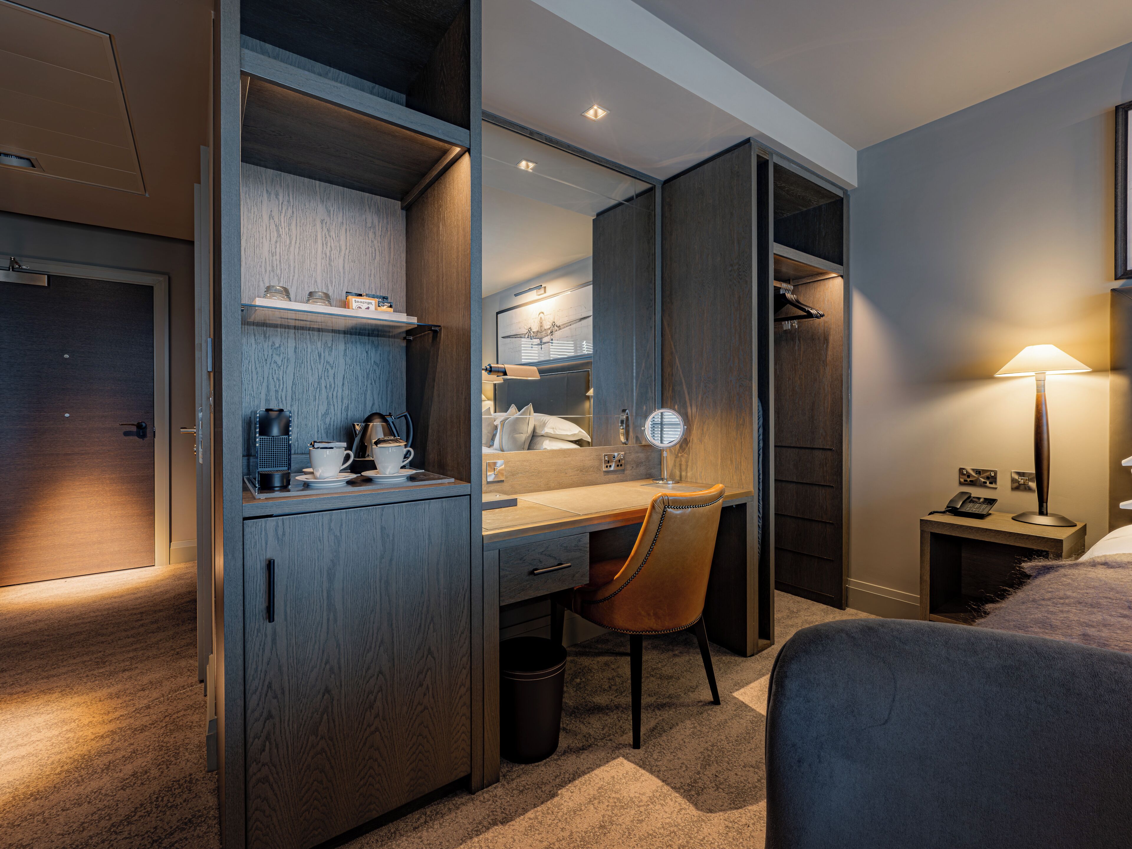 A hotel room with a built-in desk, a chair, a bed, a bedside table with a lamp, a mirror, and a wardrobe.