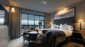 A modern hotel room with a large bed, a desk, a chair, a small table, and a view of the outdoors through large windows. - Dakota Manchester Airport (Manchester)