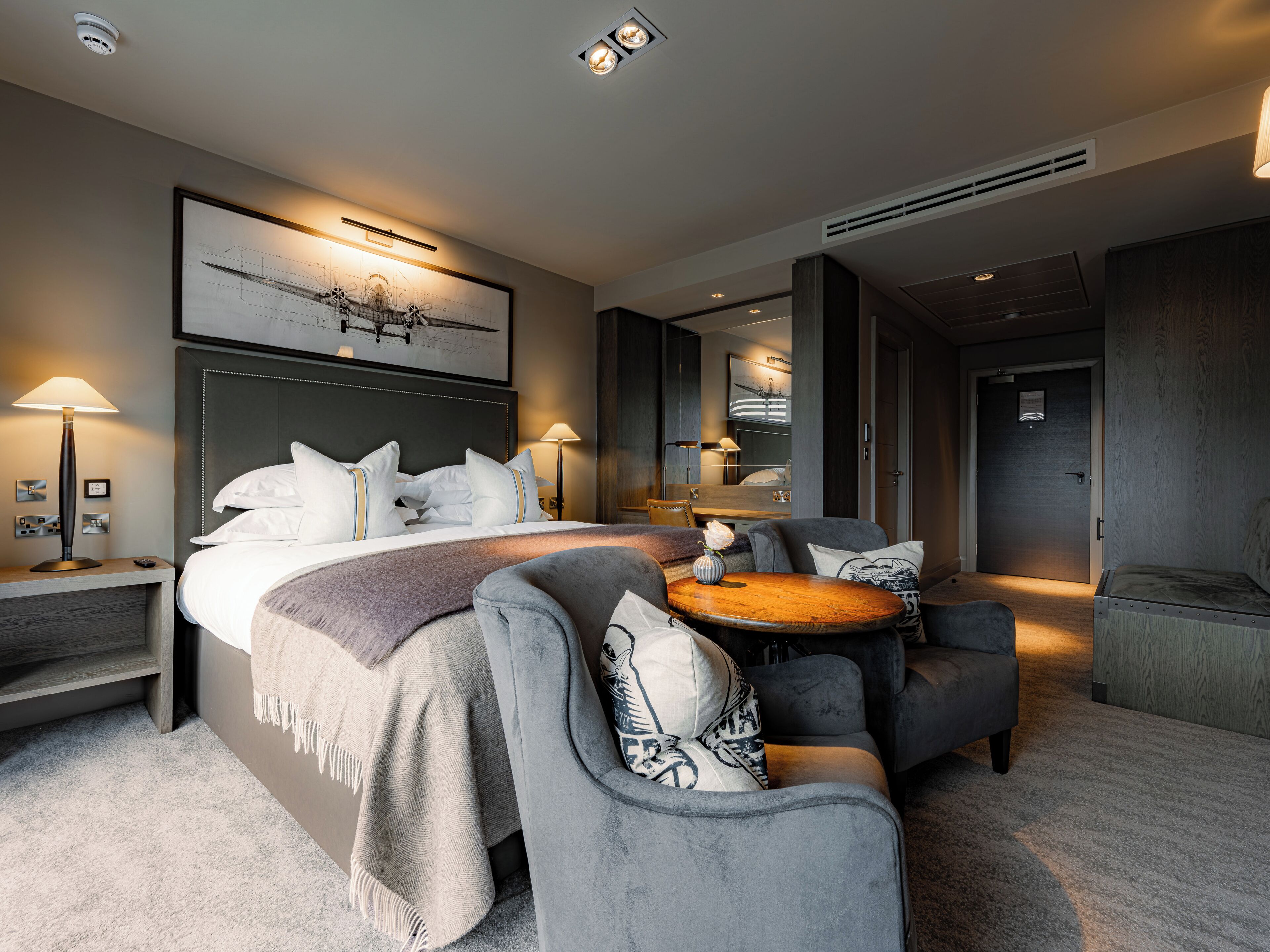 Moderni hotellihuone, jossa on suuri sänky, oleskelualue pyöreällä pöydällä sekä suuri peili.