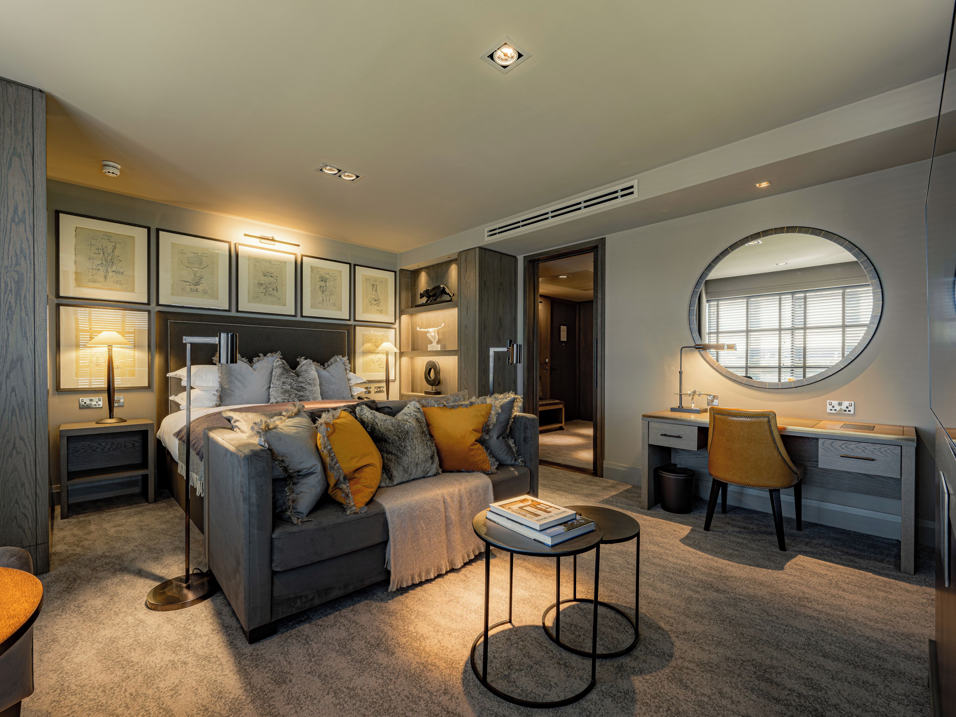A modern bedroom with a grey sofa, a round mirror, a desk, and framed artwork on the walls.