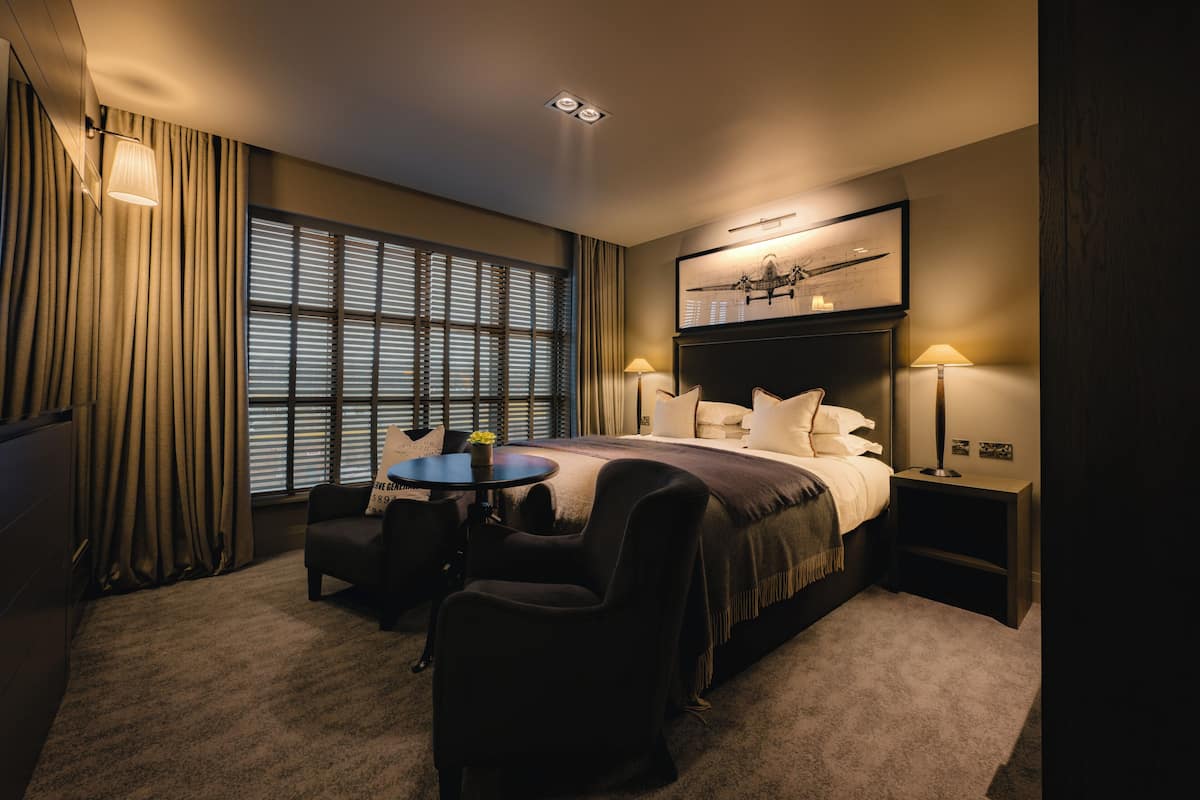 Habitación de hotel con una cama grande, un escritorio con silla, una mesita y una ventana con persianas.
