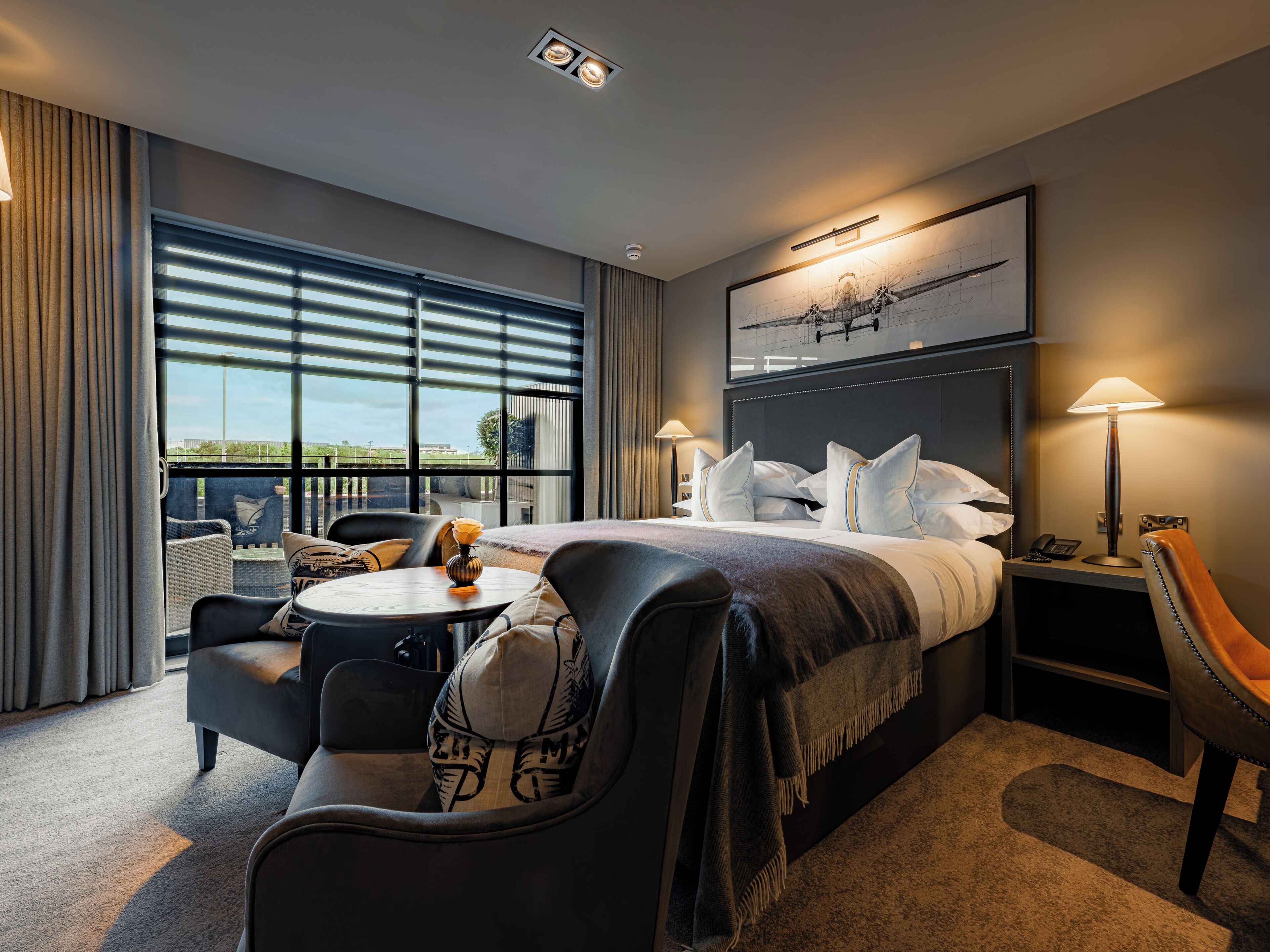 A hotel room with a large bed, a desk, a chair, a small table, and a view of the outdoors.