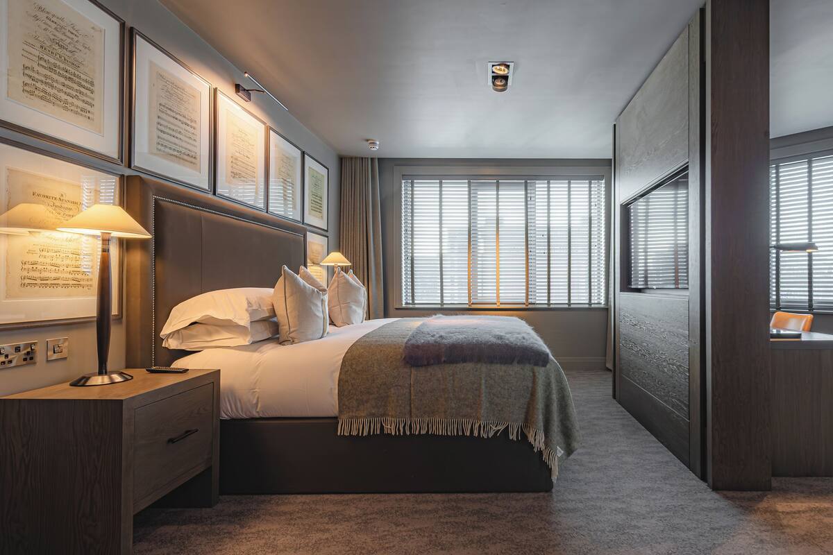 Habitación de hotel con una cama grande, una mesita de noche, una lámpara, una ventana con persianas y documentos enmarcados en la pared.
