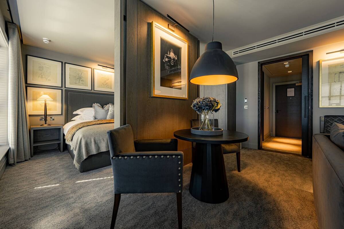 Habitación de hotel con cama, mesita de noche, lámpara, silla, mesa redonda con un florero y cuadros en la pared.