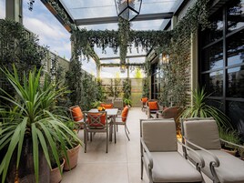A covered outdoor seating area with tables and chairs, surrounded by potted plants and a glass ceiling.