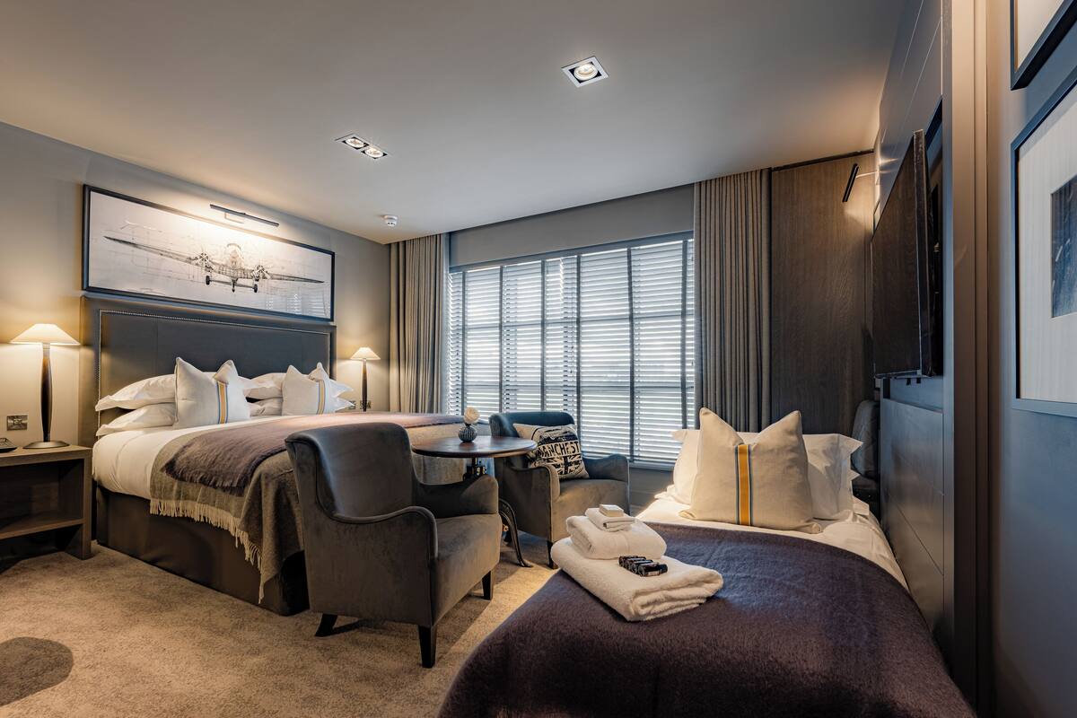 Habitación de hotel con cama, dos sillones, una mesita, escritorio, ventana con persianas y una lámpara de techo.