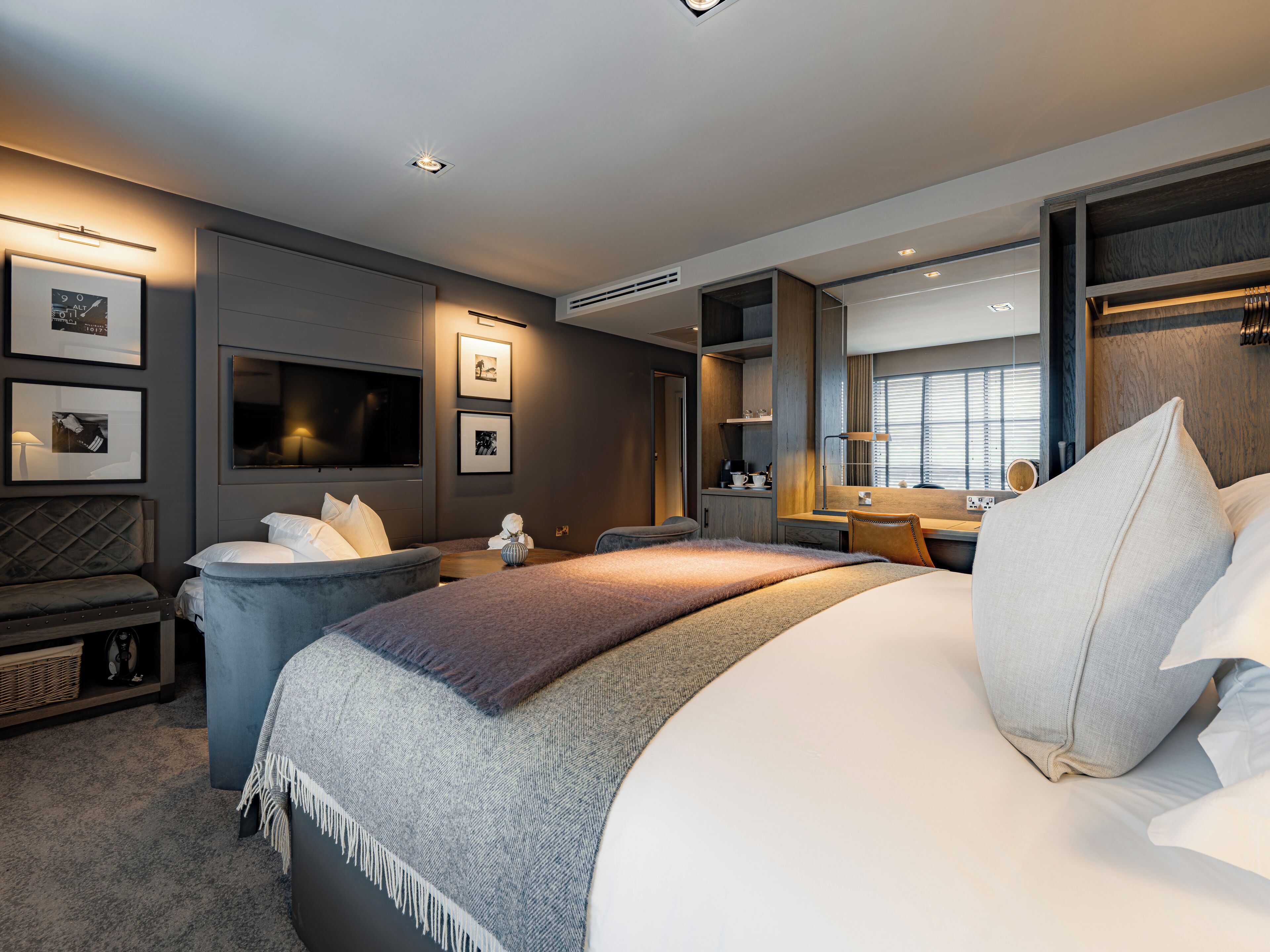 A modern hotel room with a large bed, a seating area with a sofa and armchair, a desk, and a television.