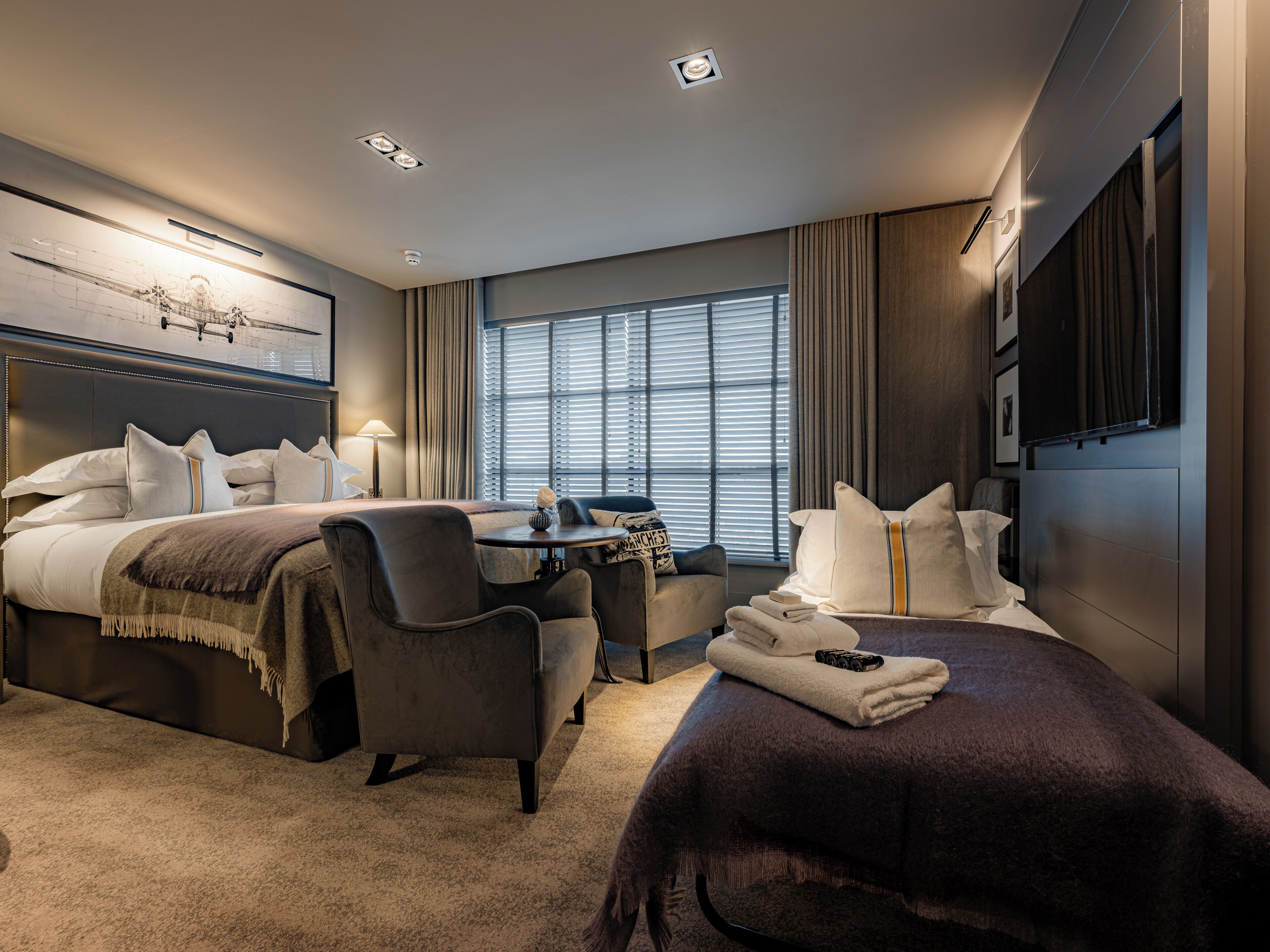 A hotel room with a king-sized bed, a sitting area with a table and chairs, a flat-screen TV, and a large window with blinds.