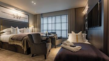 A hotel room with a king-sized bed, a sitting area with a table and chairs, a flat-screen TV, and a large window with blinds.