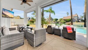 Un patio soleado con mobiliario de mimbre, un ventilador de techo y un televisor mostrando un paisaje pintoresco.