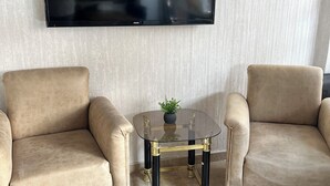 A living room with two beige armchairs, a glass coffee table with brass legs, and a flat-screen TV mounted on the wall. - Hotel KONAK (Gaziantep)