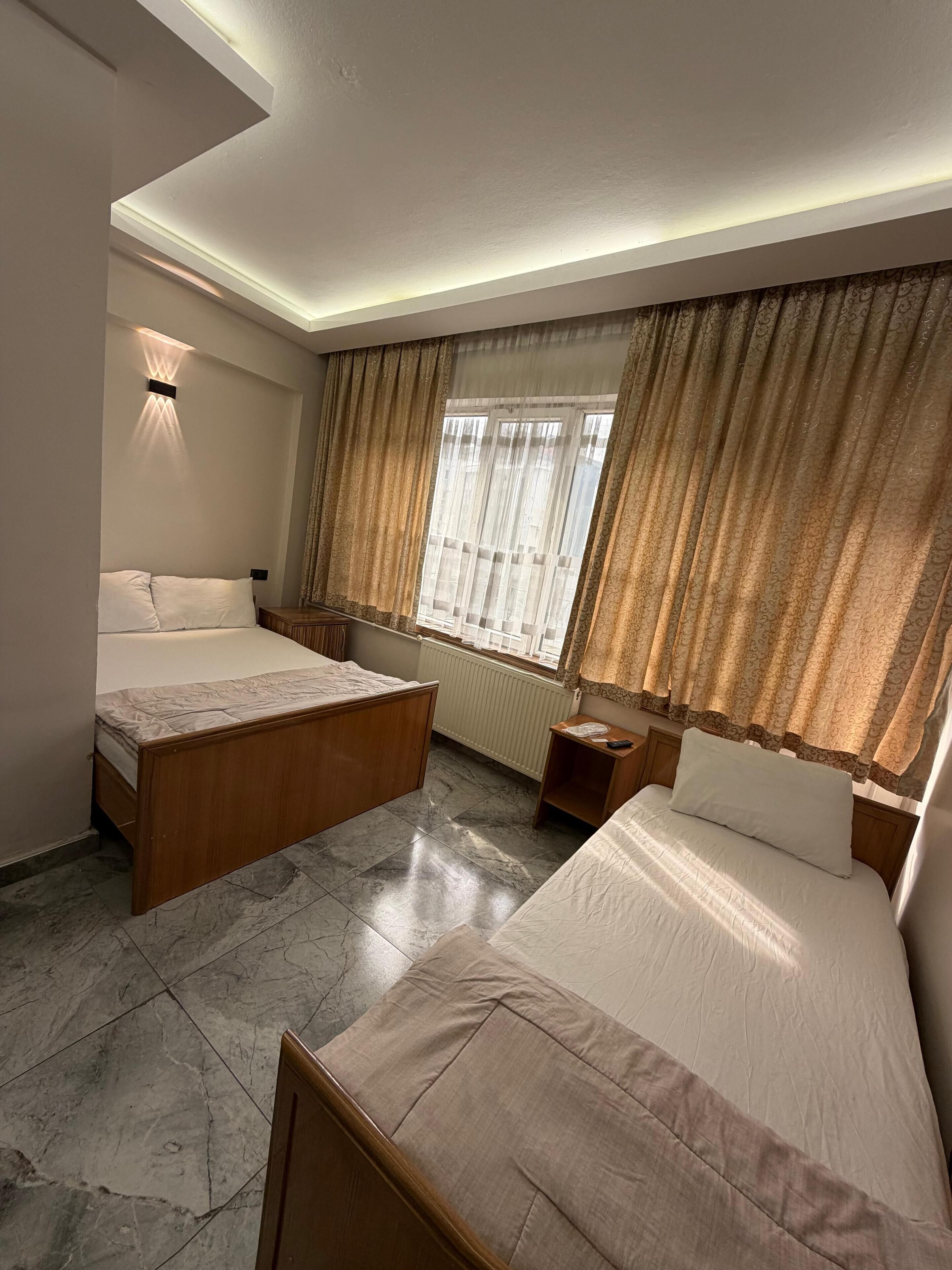 A hotel room with two beds, a wooden headboard, a bedside table, a nightstand, a window with curtains, and a wall-mounted light fixture.
