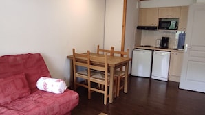 A compact living space with a wooden dining table and chairs, a red sofa, a kitchenette with a microwave and coffee maker, and a white door. - 4 pers. apartment with quiet balcony, pool access (Besse-et-Saint-Anastaise-Super Besse)