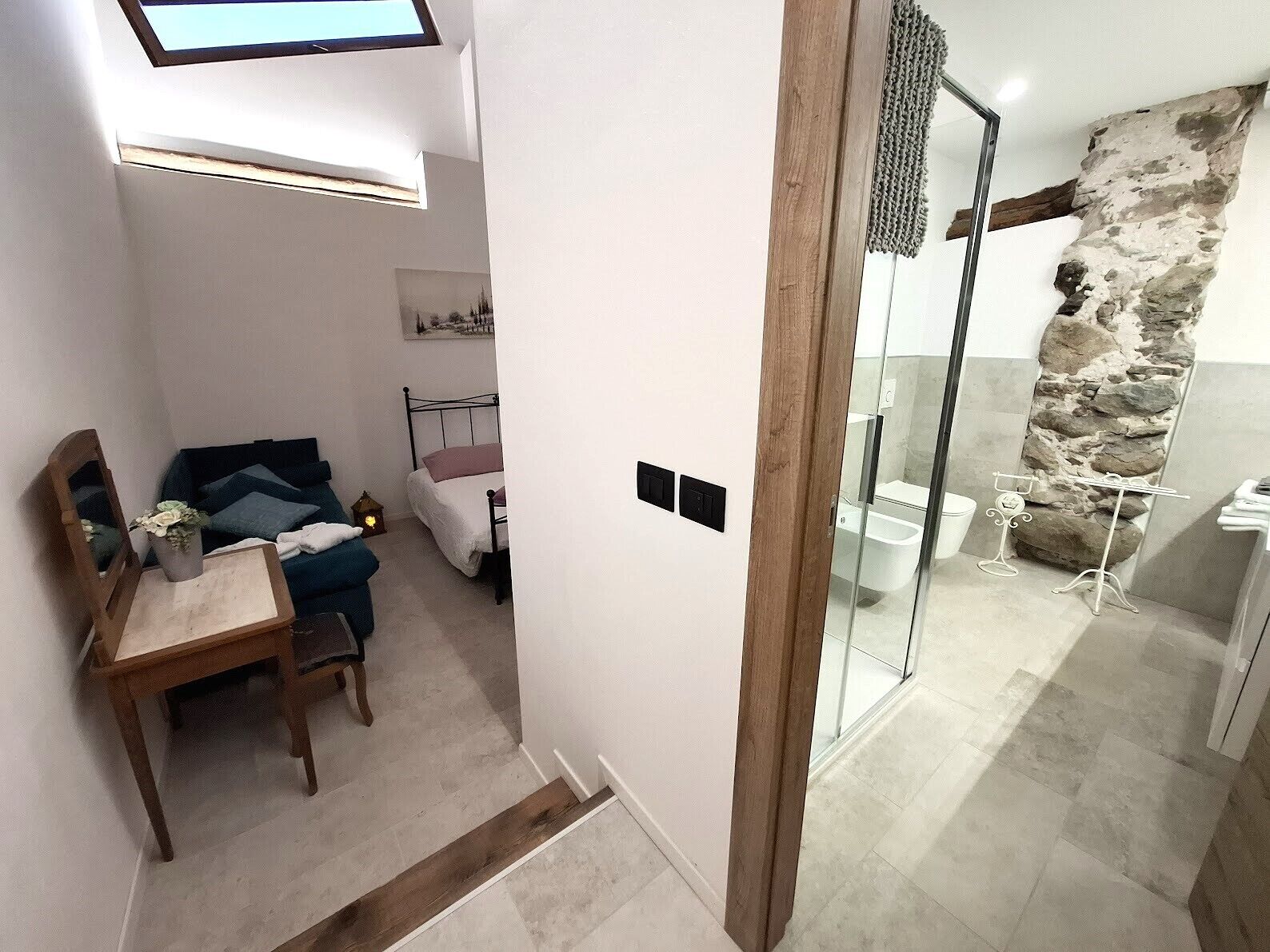 Ein modernes Schlafzimmer mit Bett, Nachttisch und kleinem Schreibtisch mit Stuhl. Es gibt ein Badezimmer mit Dusche, Toilette und einer Wand mit Steinverkleidung.
