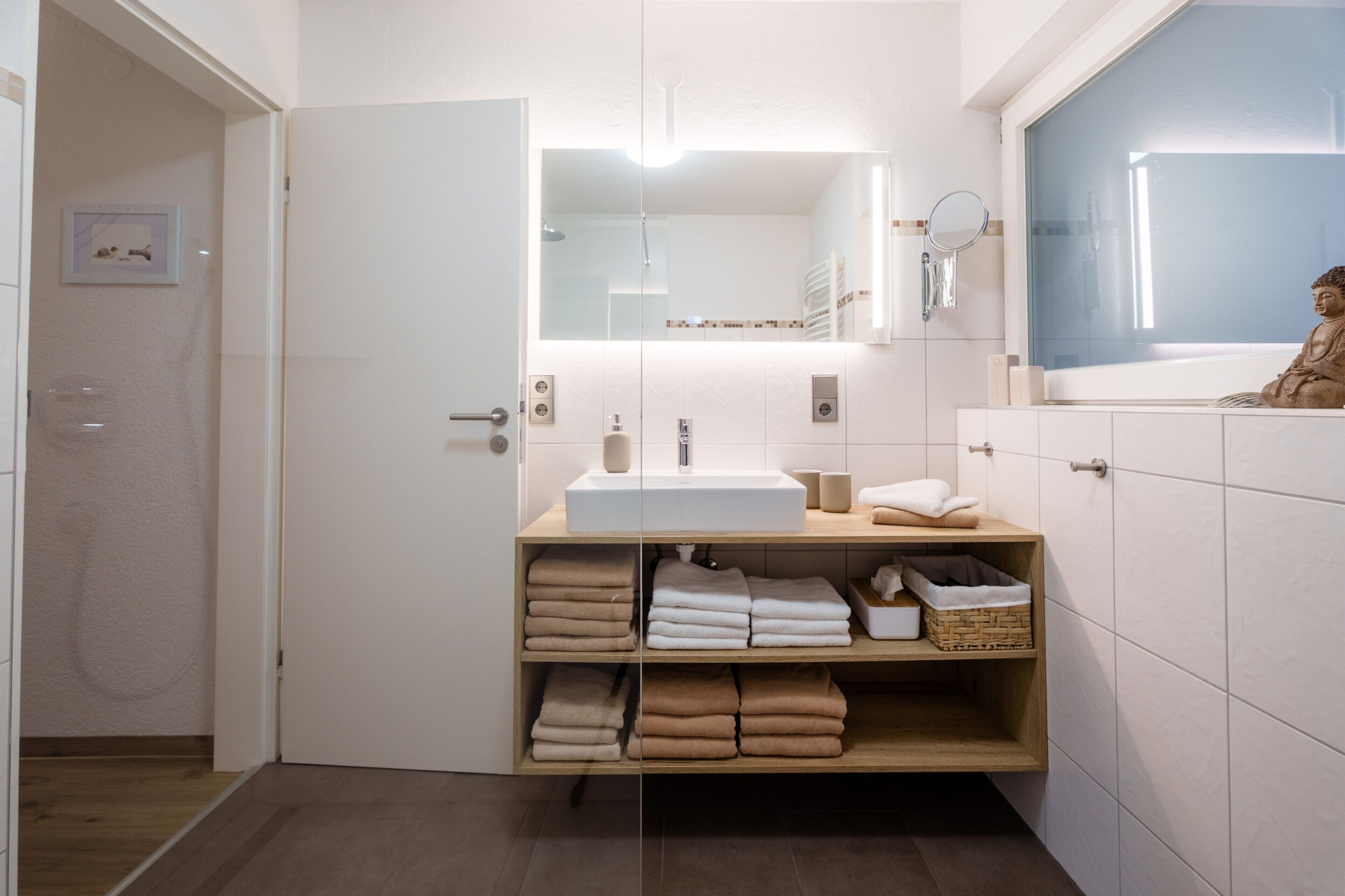 Een moderne badkamer met een witte wastafel, spiegel en planken voor handdoeken.
