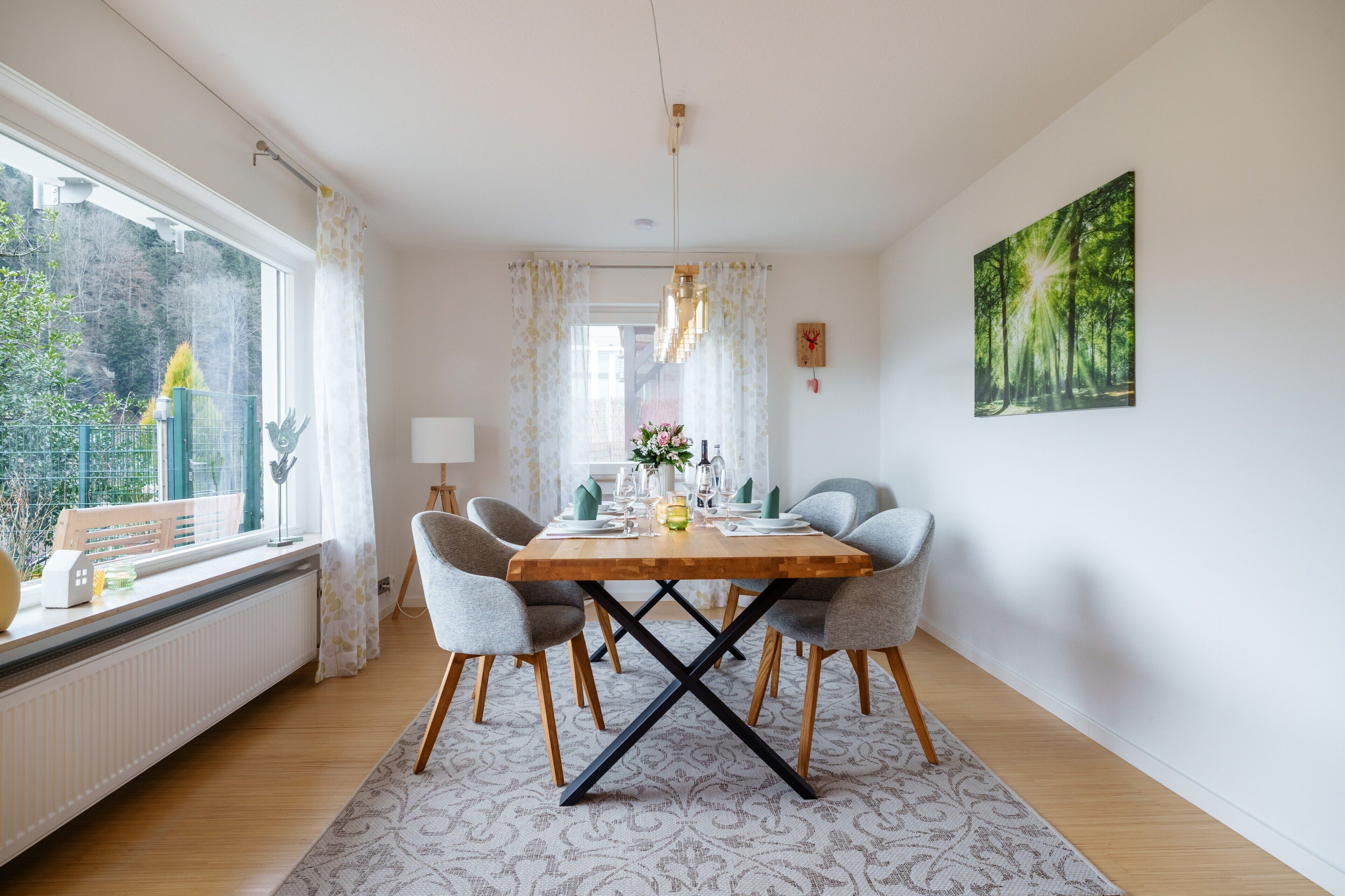 Et spiseområde med et træbord, fire grå stole, en lysekrone, en radiator og et vindue med gardiner.