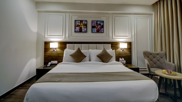 A hotel room with a large bed, two bedside lamps, a headboard with a wooden panel, a small table with fruit, and a chair. - SK PREMIUM PARK SONIPAT (Ganaur)