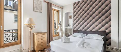 A bedroom with a large bed, wooden floor, a dresser, a lamp, and a window with curtains.