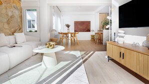 Een moderne woonkamer met een witte bank, een ronde salontafel en een houten kast. Er is een eethoek met een tafel en stoelen, en een groot schilderij aan de muur.