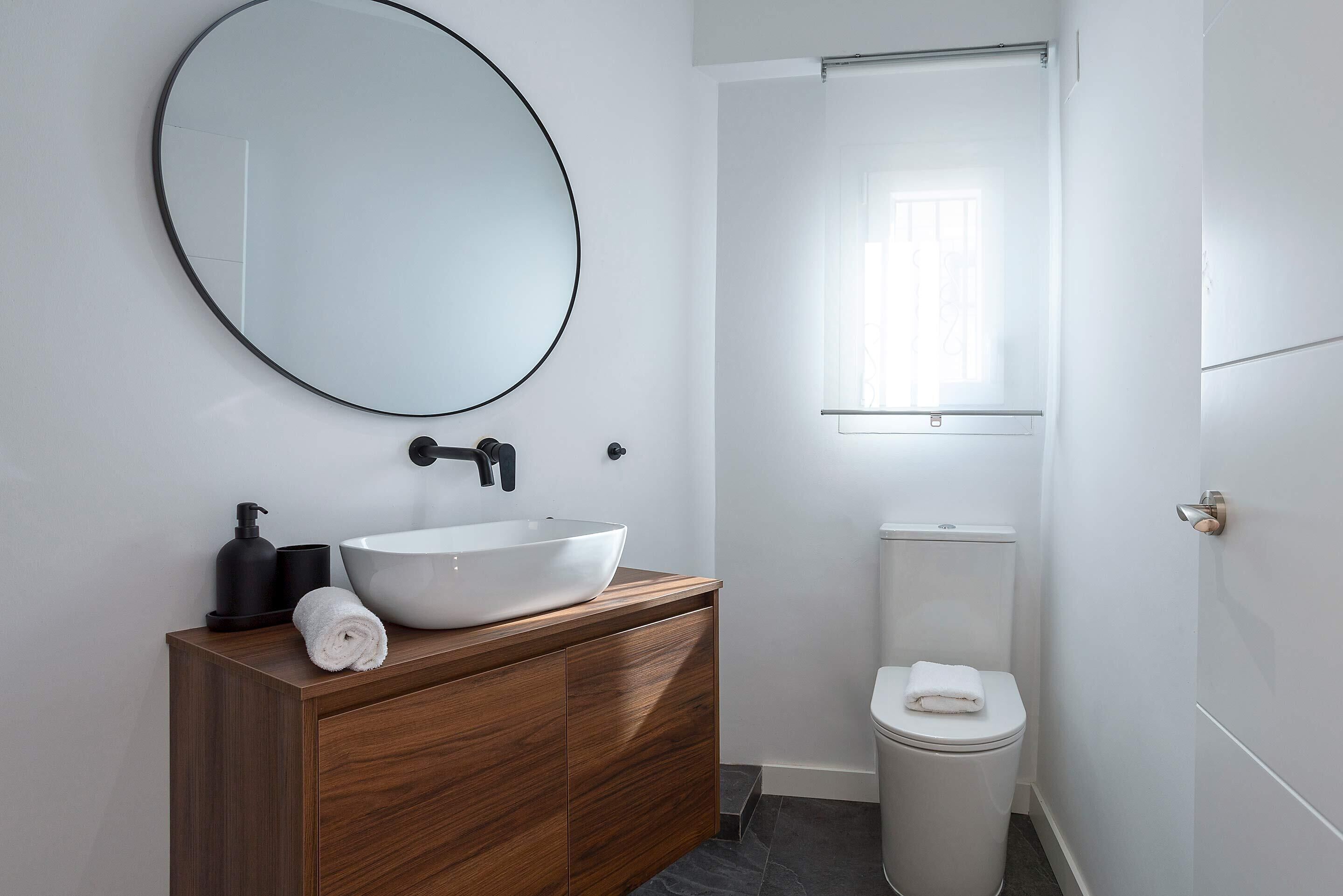 Een moderne badkamer met een ronde spiegel, een houten wastafelmeubel, een witte wasbak, een toilet en een raam voor natuurlijk licht.