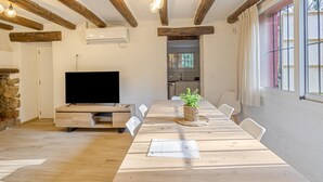 A dining area with a long wooden table, chairs, a flat-screen TV, a potted plant, and a stone wall. - Holiday Home 'La Caseta De L'Adela' with Mountain View, Wi-Fi and Air Conditioning (Arbúcies)