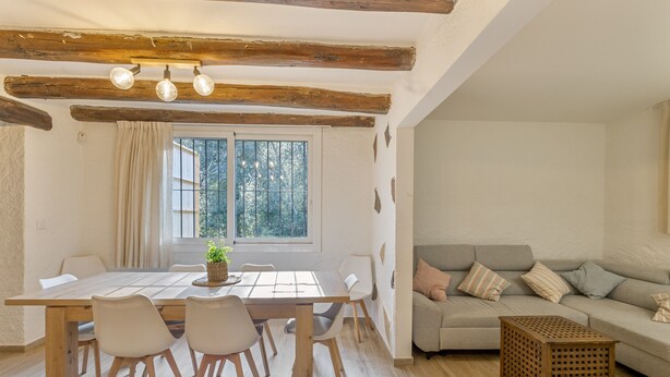 A dining area with a wooden table, chairs, a plant, and a window with curtains. - Holiday Home 'La Caseta De L'Adela' with Mountain View, Wi-Fi and Air Conditioning (Arbúcies)