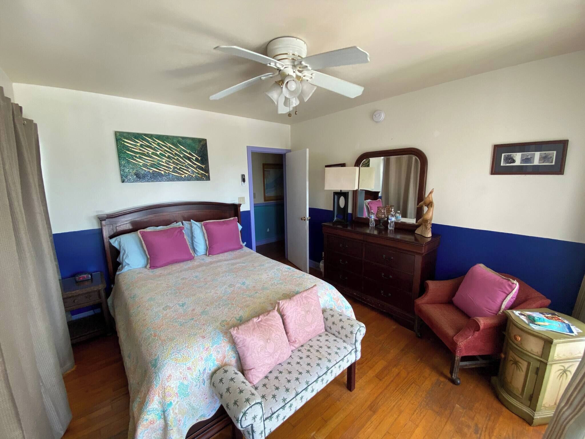 A hotel room with a bed, a dresser, a chair, a ceiling fan, a mirror, and a painting on the wall.