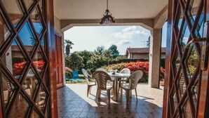 Een open terras met een tafel en stoelen, een hanglamp en uitzicht op een tuin met rode bloemen en bomen.