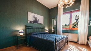 Een slaapkamer met een bed, twee bedlampen, een raam met gordijnen, een radiator en een aan de muur hangend schilderij.