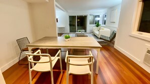 Uma sala de estar moderna com piso de madeira, uma mesa de jantar com quatro cadeiras brancas, um sofá e uma pequena planta sobre a mesa.