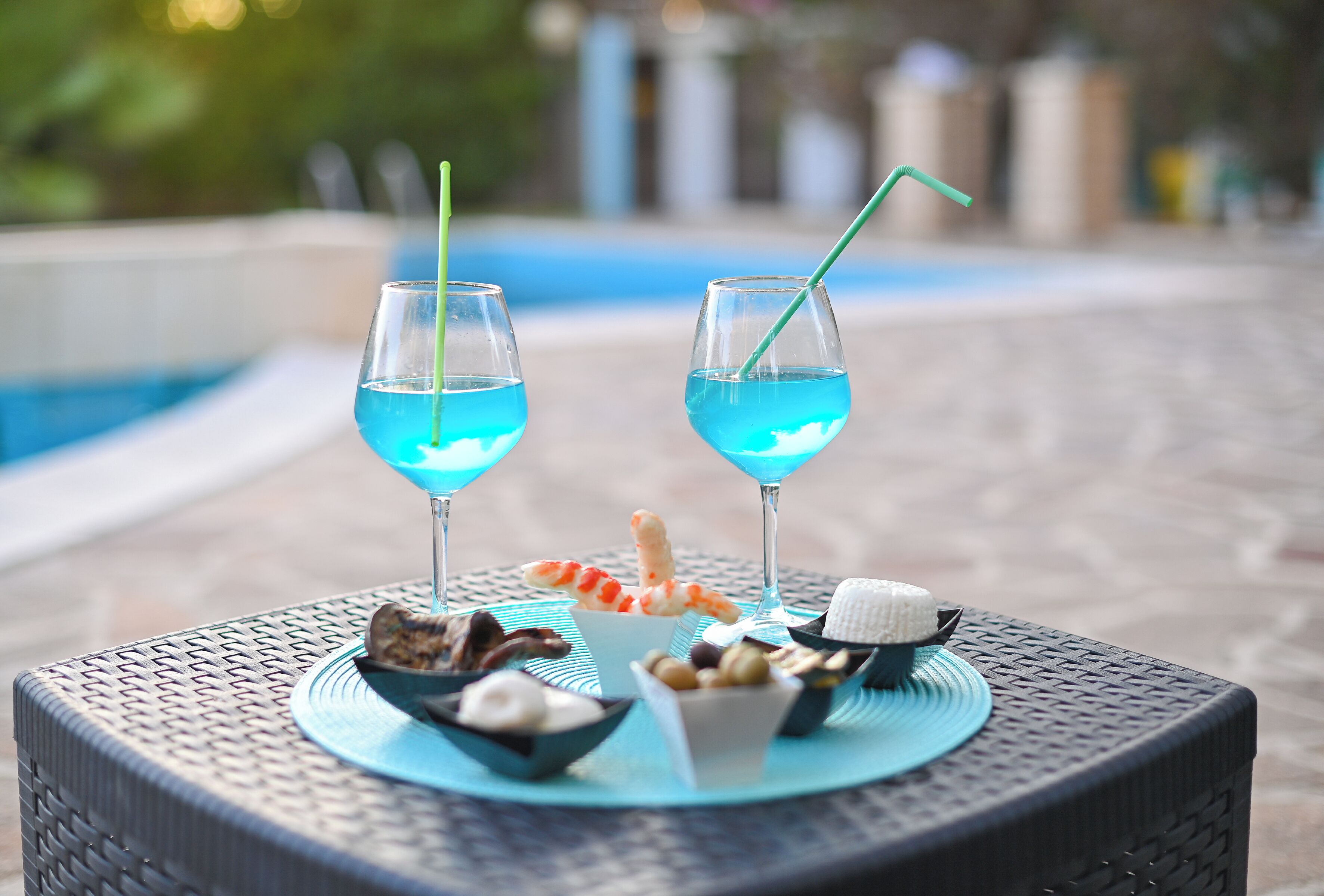 Dos vasos de cóctel azul con pajitas, un cuenco de frutos secos variados, un pequeño plato de helado y un camarón sobre una mesa redonda junto a una piscina.