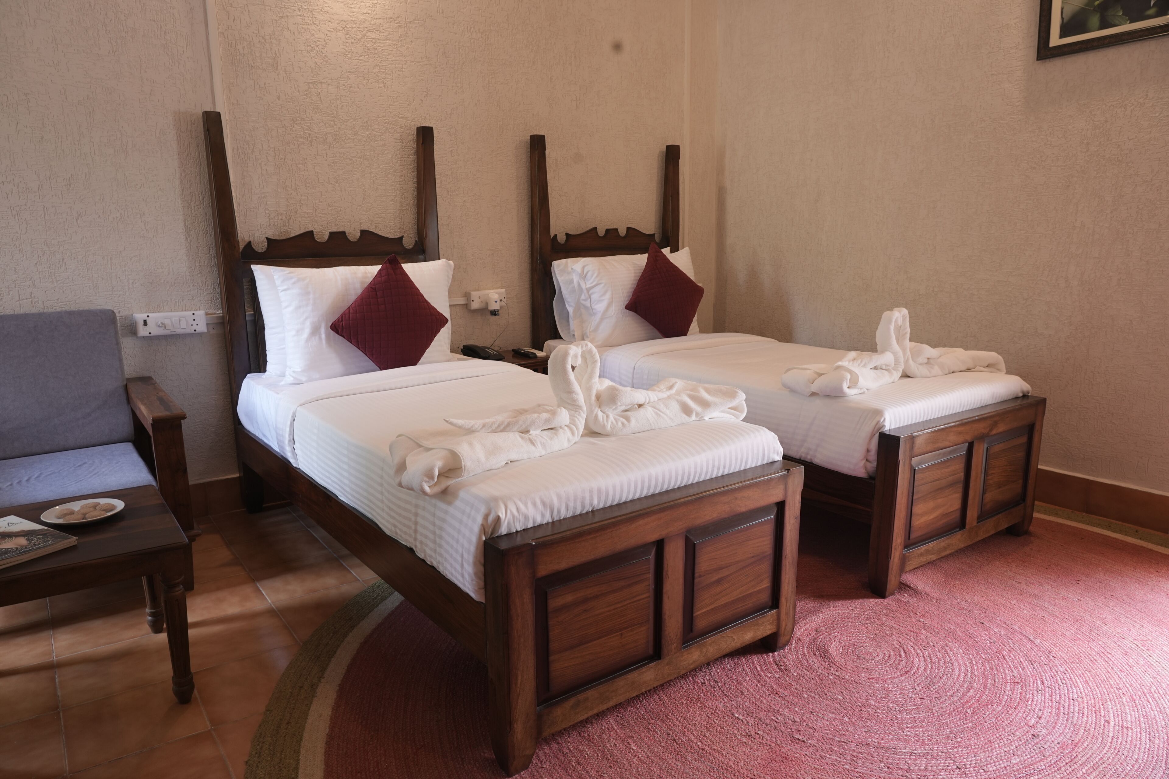 Habitación de hotel con dos camas de madera, cada una con sábanas blancas y almohadas rojas. Hay una mesita con un plato y un libro, y una silla a la izquierda.