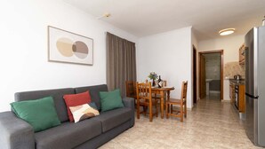 Ein Wohnzimmer mit grauem Sofa, grünen und roten Kissen, einem Holzesstisch mit Stühlen und einer Küchenzeile inklusive Kühlschrank.