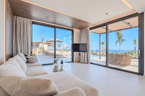 A modern living room with a sofa, a coffee table, and a TV. There are large windows offering a view of a pool and palm trees. - Nysa Luxury villa 3,Private pool,Near Hersonissos (Heraklion)