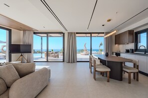 A modern living room with a sofa, a TV, a dining area with a table and chairs, and a kitchen with a large window. - Nysa Luxury villa 4, Private pool, Near Hersonissos (Heraklion)