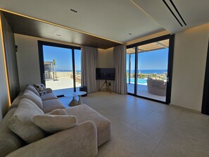 A modern living room with a large sofa, a flat-screen TV, and a view of the beach and ocean through large glass doors. - Nysa Luxury villa 4, Private pool, Near Hersonissos (Heraklion)